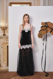 Woman in a black evening gown with white lace details standing next to a horse head sculpture in an elegant interior setting.