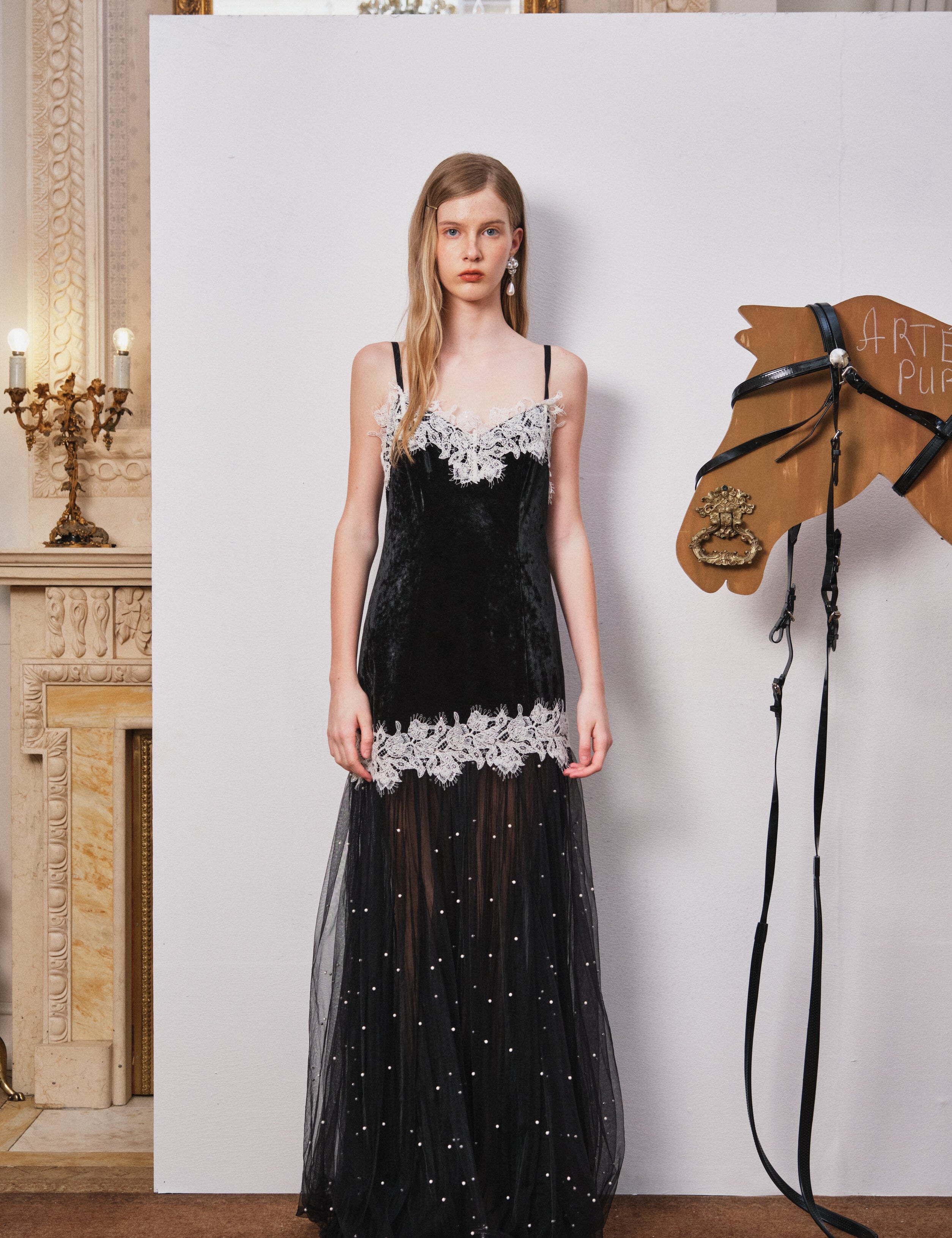 Woman in a black evening gown with white lace details standing next to a horse head sculpture in an elegant interior setting.