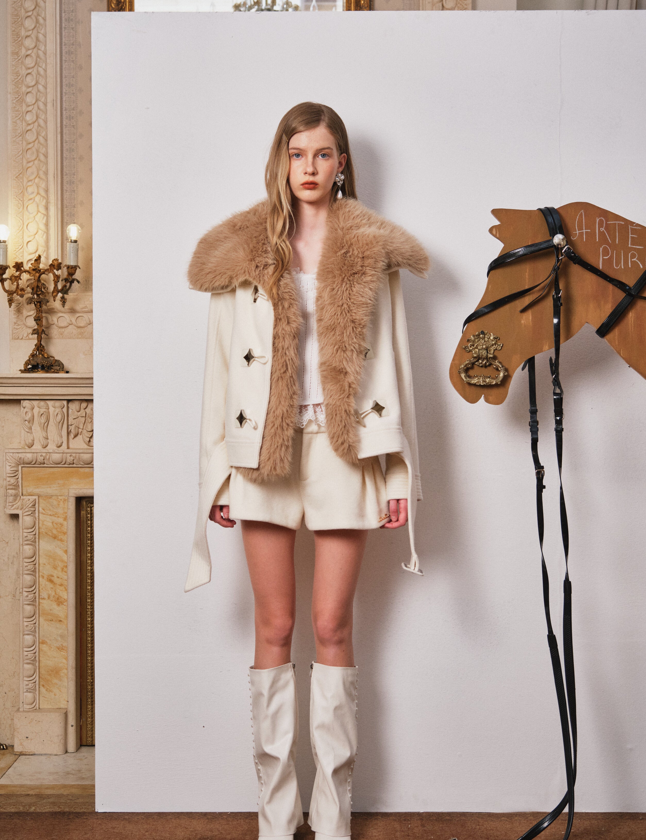 Woman in a fur-trimmed coat standing next to a wooden horse figure in an elegant room.