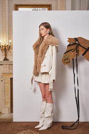 Woman in a fur vest and white boots standing next to a horse head sculpture in an elegant interior setting.