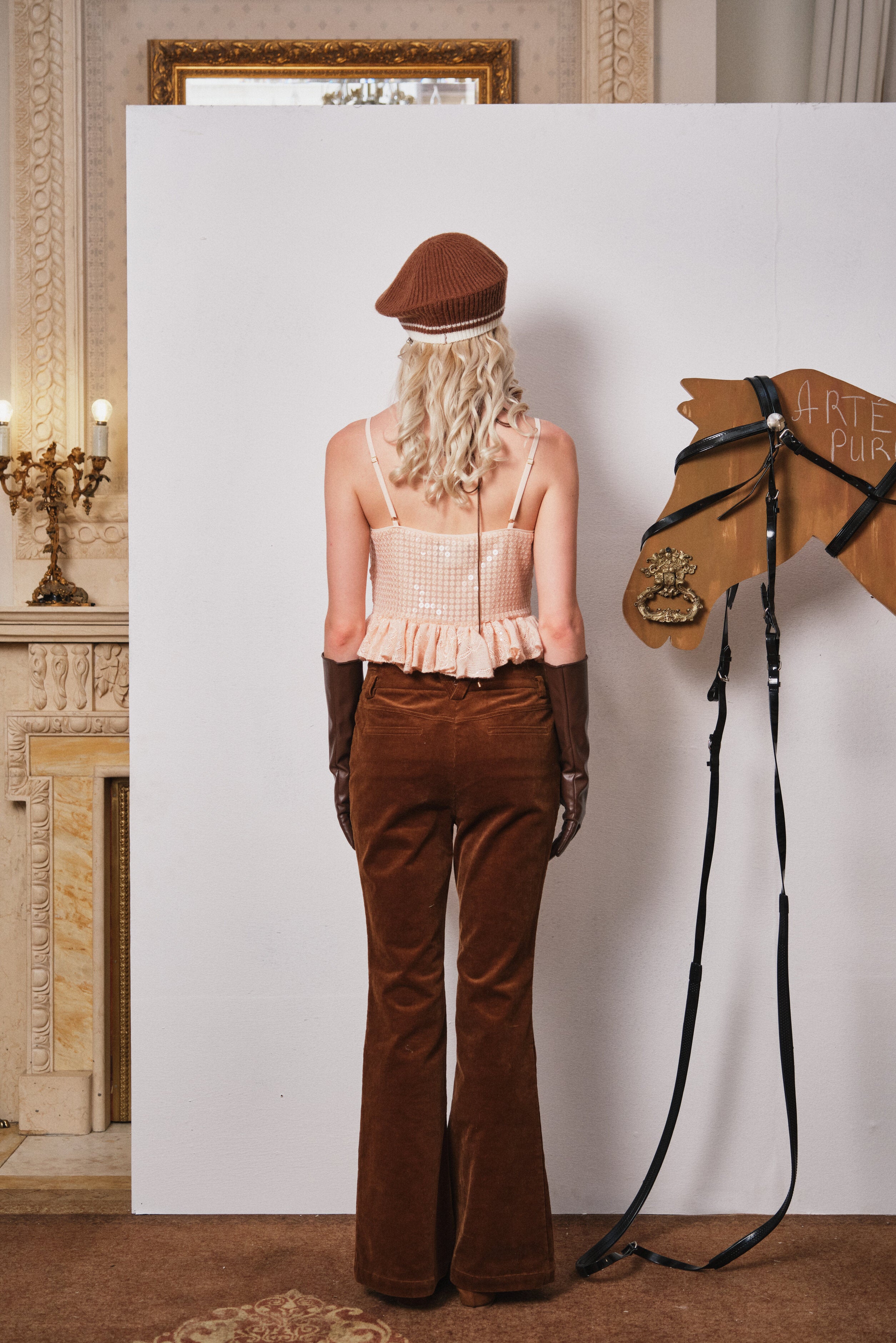 Person in vintage outfit with brown pants and hat standing next to a horse head sculpture indoors.